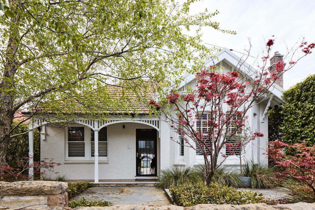 Single-story white house with a tiled roof, front porch, and a garden with green and red-leaved trees.