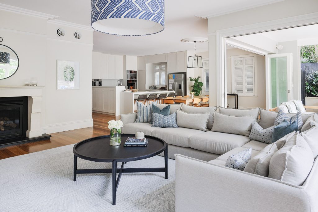 Modern living room featuring a light sectional sofa, round black coffee table, fireplace, open kitchen and dining area in the background, and large windows filling the space with natural light.
