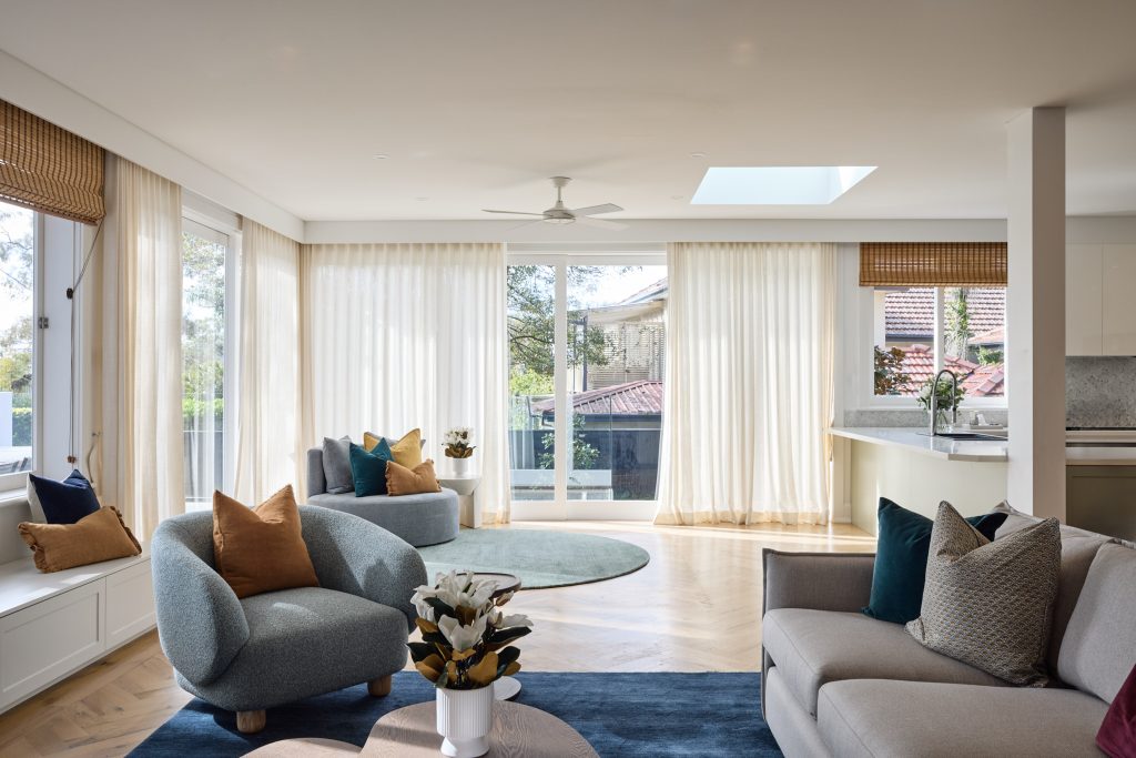 Modern living room with large windows, sheer curtains, cushioned chairs, a round rug, and a coffee table with flowers; kitchen visible in the background for an inviting Northbridge space.