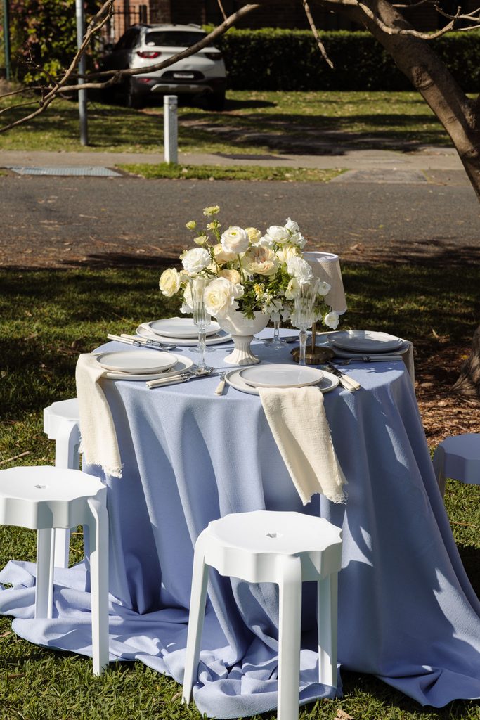 A round table set for four with a light blue tablecloth, white floral centerpiece, and four white stools placed on an Emma Blomfield rug on a grassy lawn.