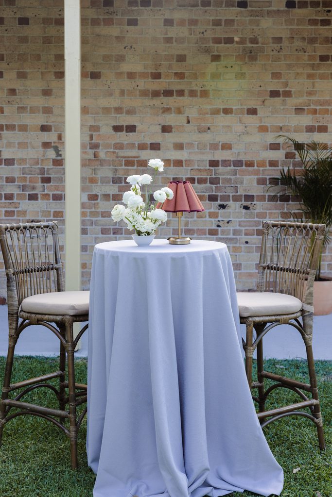 A high-top table with a light purple tablecloth and Emma Blomfield rug collection, two rattan bar stools, white flowers, and a pleated lamp set against a brick wall.
