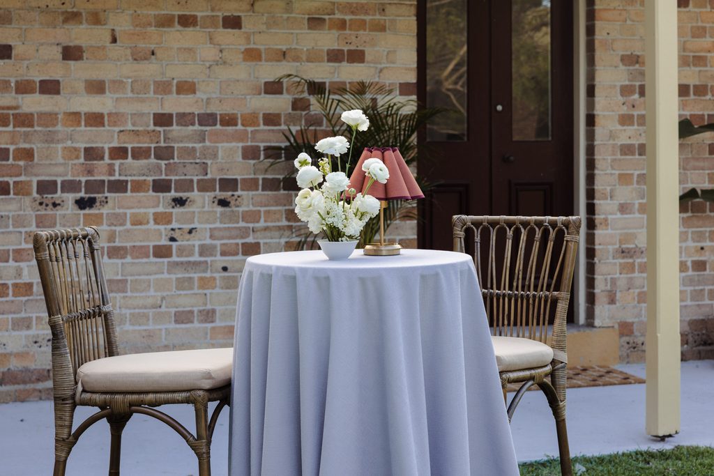 A round table with a grey cloth, a vase of white flowers, and a small lamp sit on an Emma Blomfield rug between two wicker chairs on a patio.