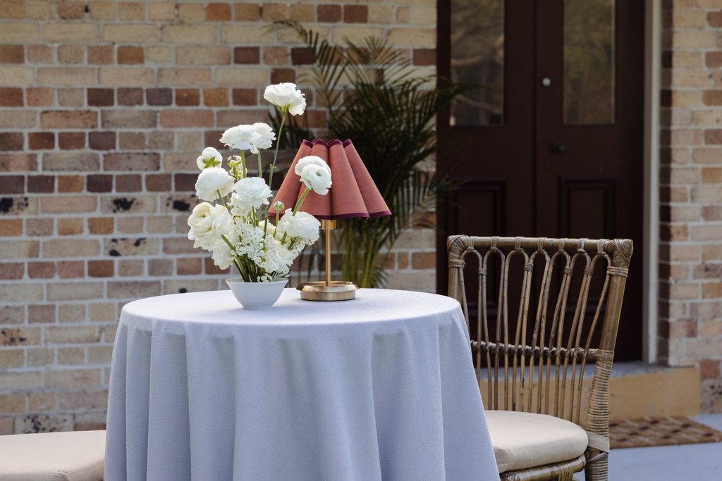A round table with a grey tablecloth sits on an Emma Blomfield rug next to a wicker chair, topped with a white flower arrangement and a small table lamp.