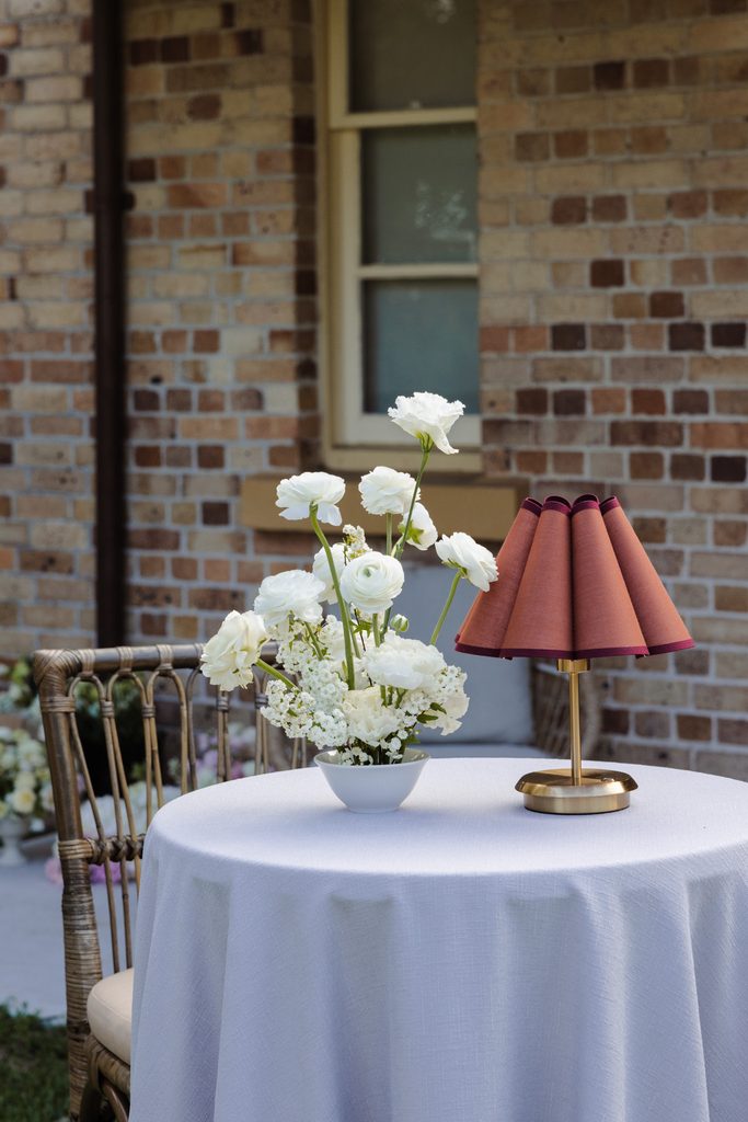 A small round table holds a white bowl of white flowers and a small lamp with a pleated maroon shade, sitting on an Emma Blomfield rug.