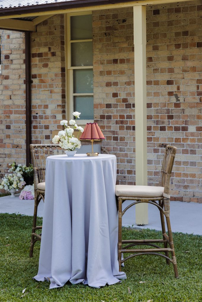 A small round table with a grey cloth sits on an Emma Blomfield rug between two rattan chairs on a lawn, accented by flowers and a lamp.