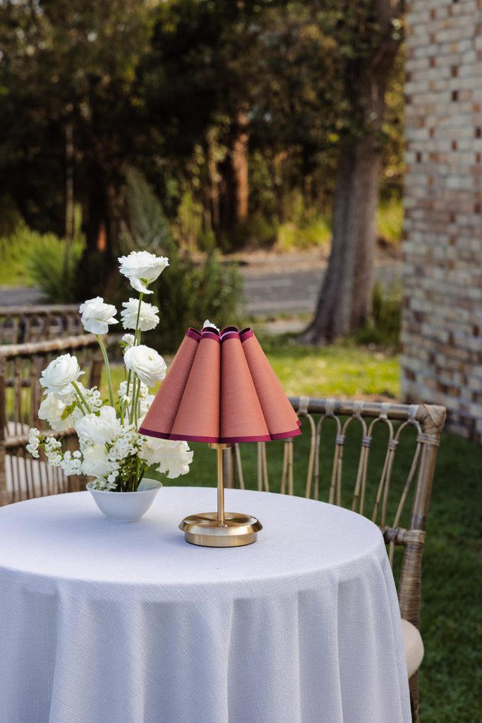 A vase of white flowers and a lamp with a pleated rust-colored shade sit on a linen-covered table atop an Emma Blomfield rug.