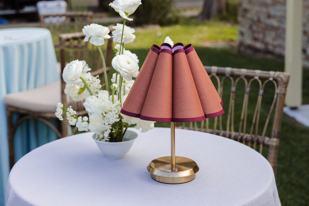 A small cordless lamp with a pleated rust-colored shade and a vase of white flowers sit on an Emma Blomfield rug collection table.