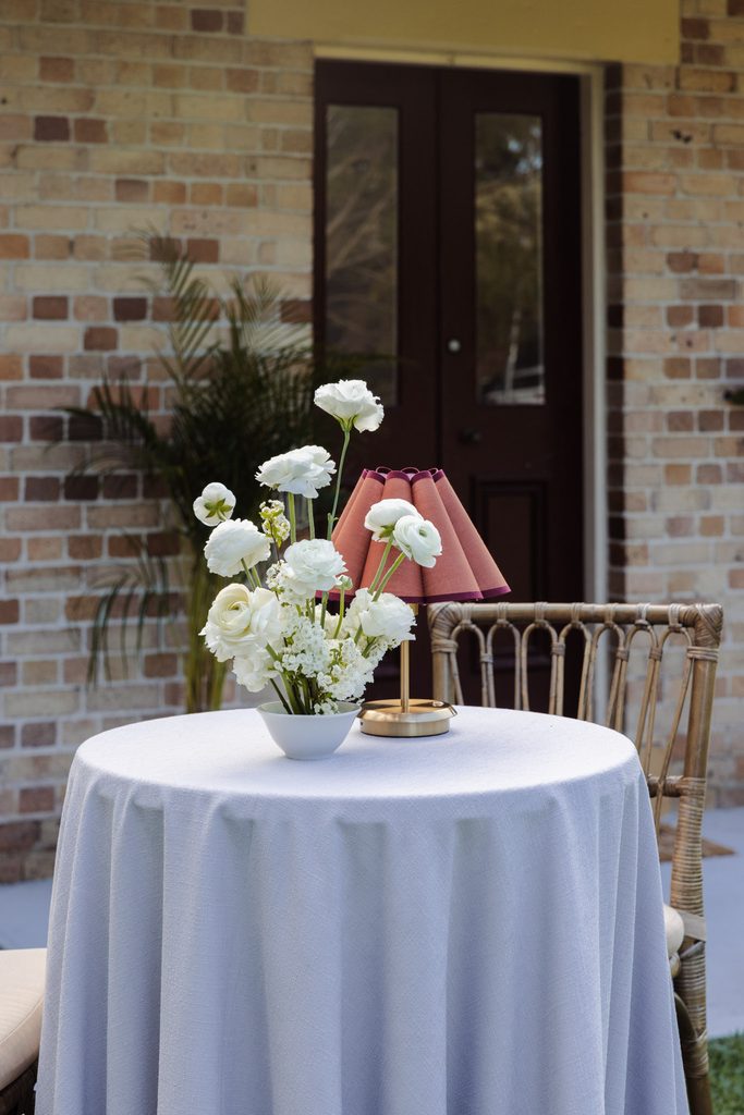 A small round table covered with a white cloth, set with a vase of white flowers and a lamp atop an Emma Blomfield rug.