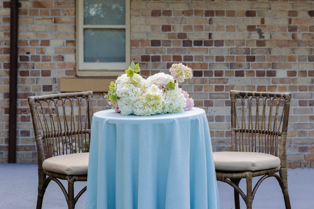 A small table with a light blue tablecloth and a hydrangea centerpiece, flanked by two rattan chairs and an Emma Blomfield rug in front of a brick wall.