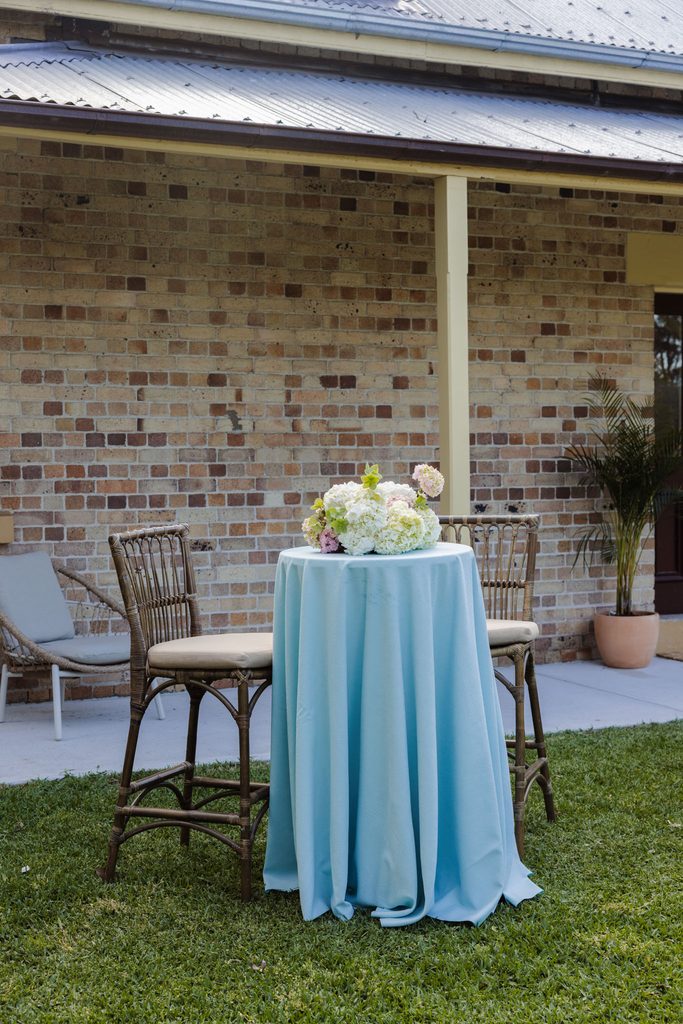 Emma Blomfield sits at a tall cocktail table with a light blue tablecloth and a floral centerpiece, accompanied by two rattan chairs on an Emma Blomfield rug on the lawn.