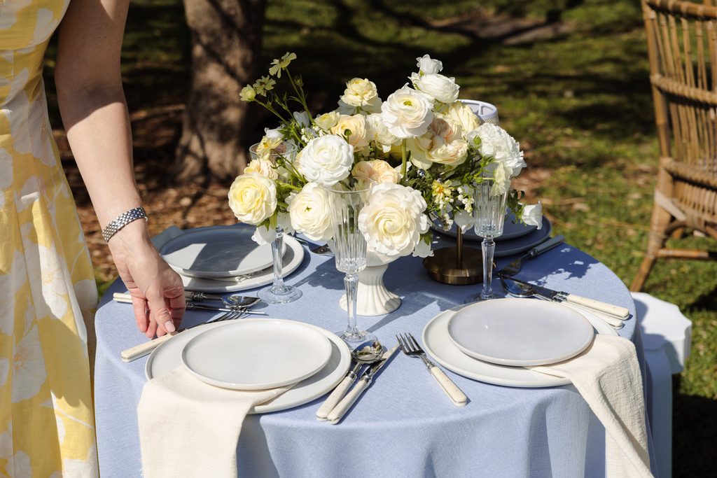 Emma Blomfield sets an outdoor table with a floral centerpiece, plates, and cutlery on a blue tablecloth atop an Emma Blomfield rug.