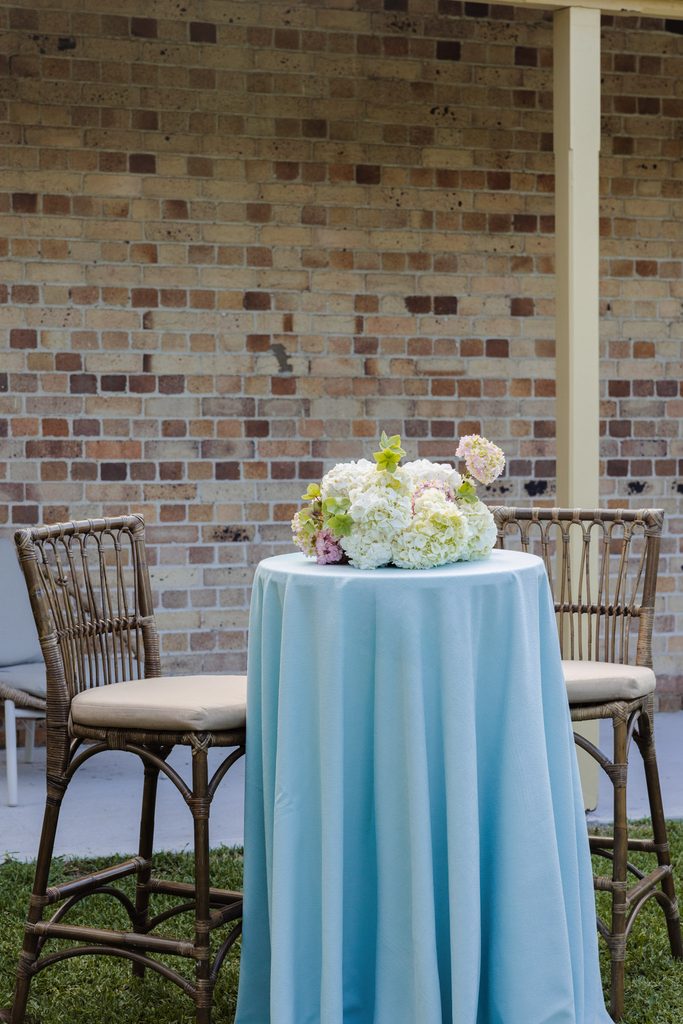 A tall bistro table with a light blue tablecloth and a floral centerpiece stands on an Emma Blomfield rug between two wicker chairs in front of a brick wall.