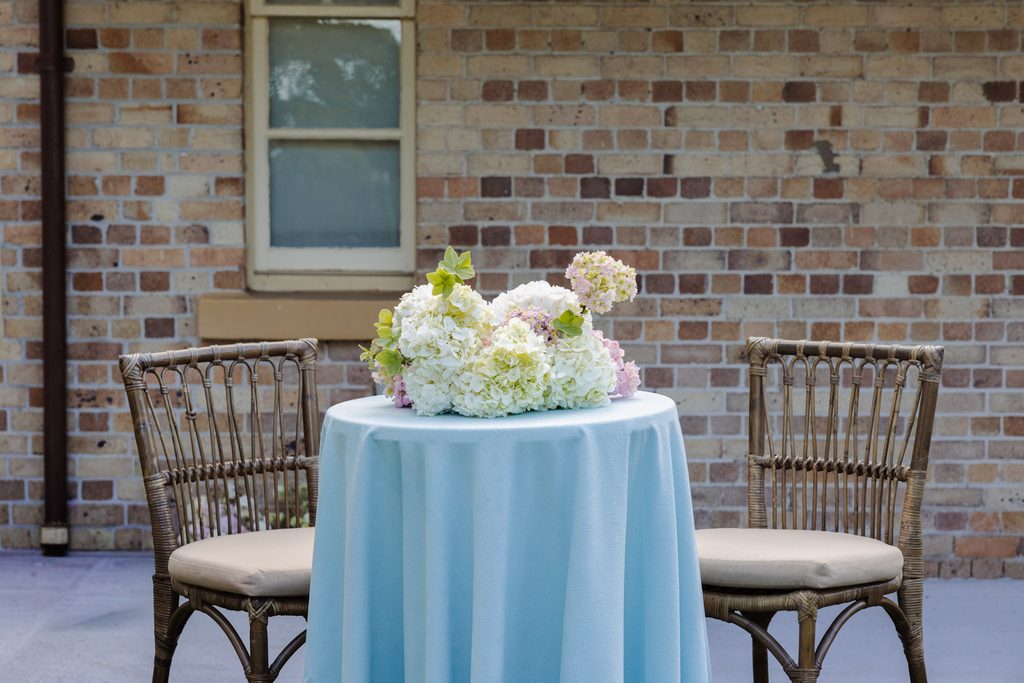 Two rattan chairs from the Emma Blomfield rug collection flank a light blue-covered table with a hydrangea centerpiece against a brick wall.