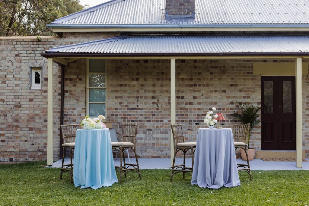 Two high-top tables with blue and grey Emma Blomfield rug collection tablecloths, floral arrangements, and chairs sit on a lawn in front of a brick house.