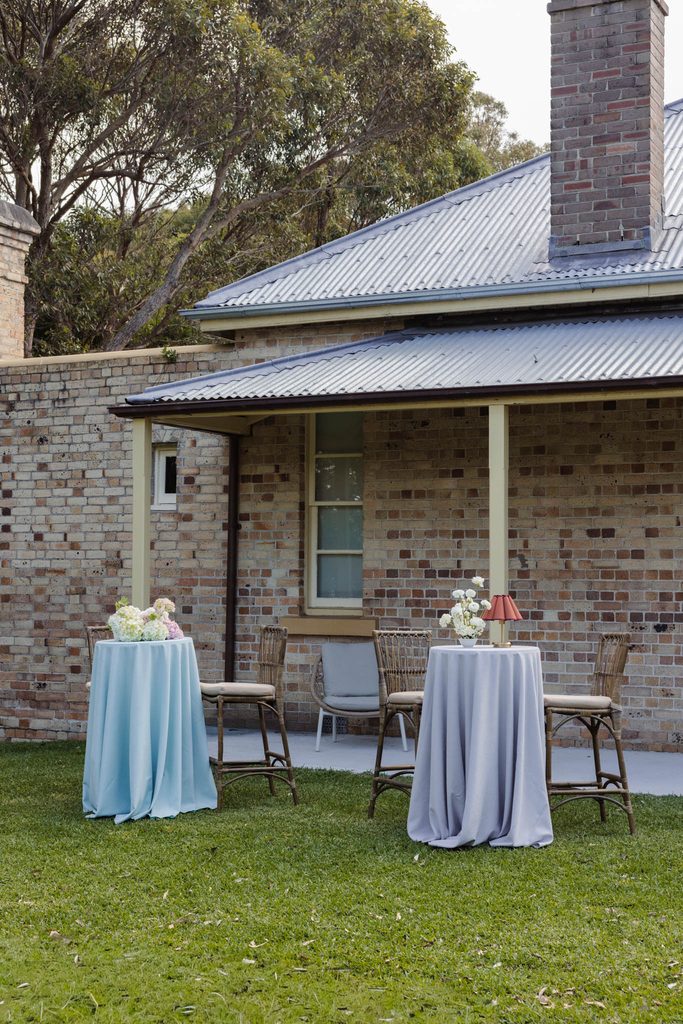 Two cocktail tables with floor-length cloths from the Emma Blomfield rug collection and floral centerpieces are set on a lawn in front of a brick house.