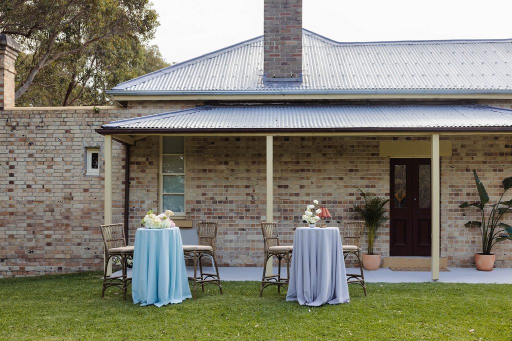 Emma Blomfield stands by two tall bistro tables with floor-length blue and grey tablecloths from the Emma Blomfield rug collection set up on a lawn in front of a brick house.