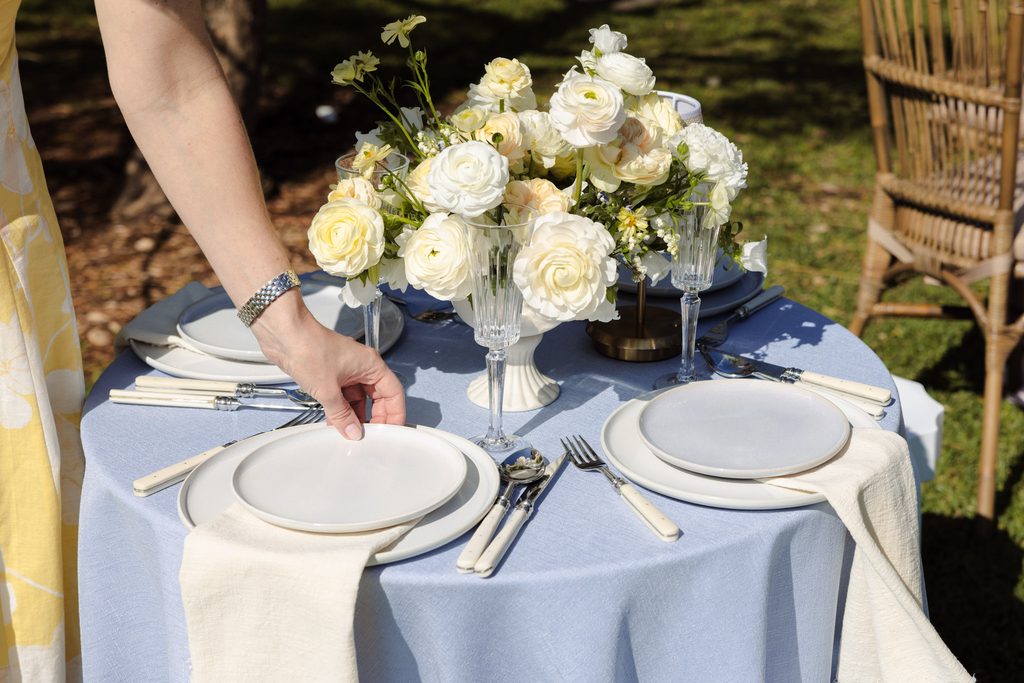Emma Blomfield sets a plate on a round table atop an Emma Blomfield rug, featuring a cream flower centerpiece.