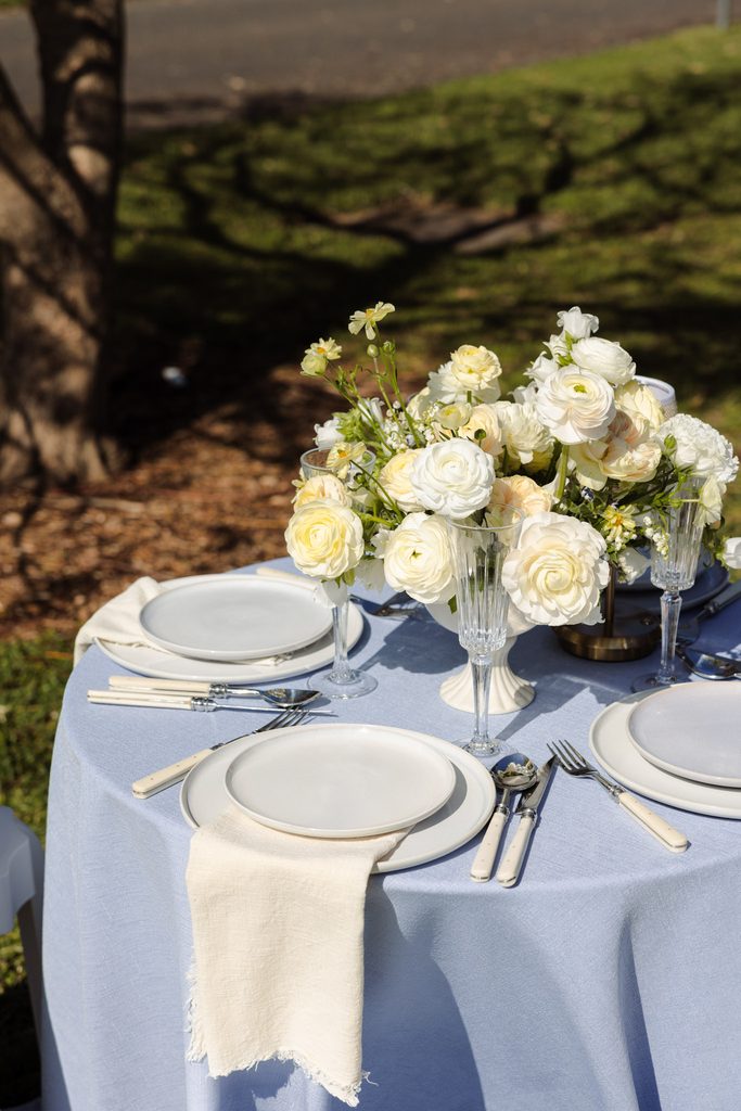 A round table with a blue tablecloth, set on an Emma Blomfield rug with white plates, flatware, and a central arrangement of white flowers.