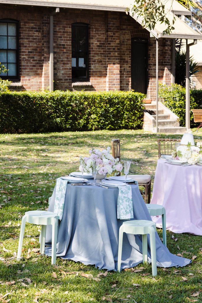 Two tables set for outdoor dining on an Emma Blomfield rug collection, positioned on a lawn in front of a brick building.