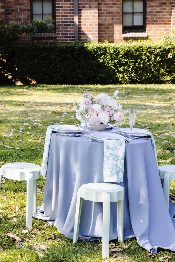 A round table set for two with a floral centerpiece and sage green stools sits on an Emma Blomfield rug collection piece atop a grassy lawn.