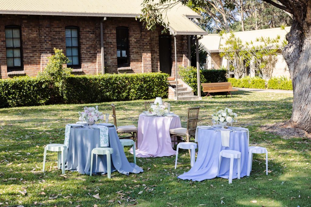 Emma Blomfield sits at one of three small tables draped in blue, pink, and lilac tablecloths from her rug collection, set for an outdoor meal on a lawn in front of a brick building.