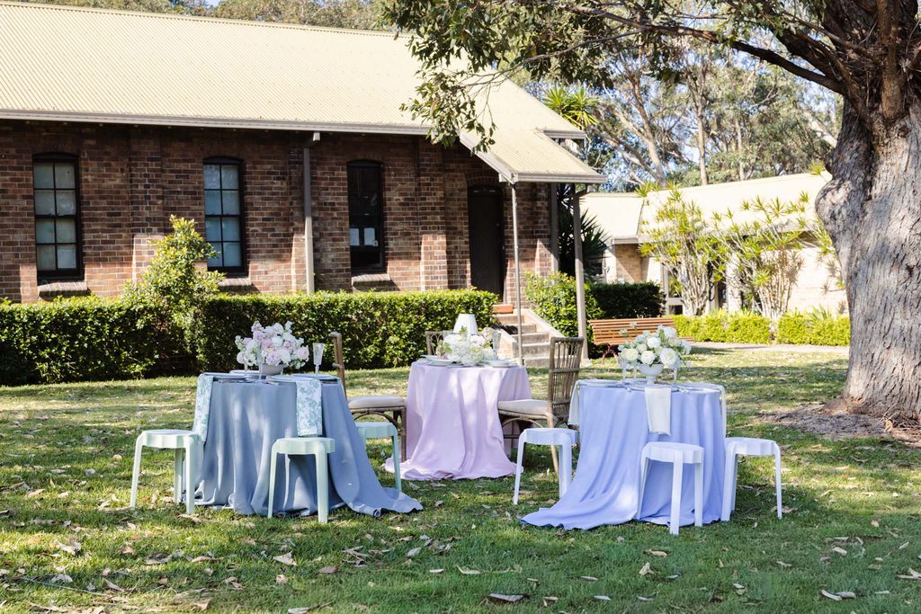 Three tables set with tablecloths and floral arrangements on a lawn in front of a brick building from the Emma Blomfield rug collection.