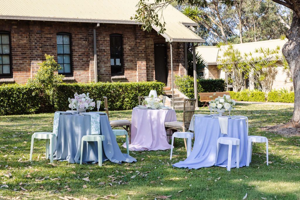 Three tables set for outdoor dining with floral centerpieces and colorful Emma Blomfield rug collection linens on a grassy lawn in front of a brick building.