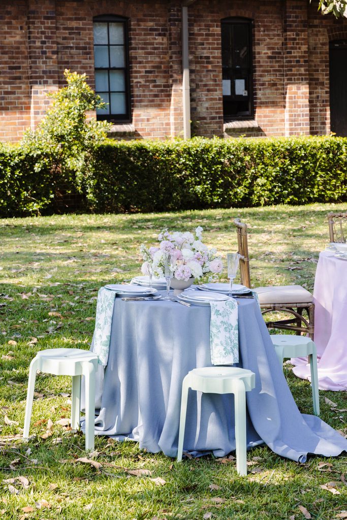 A table set for dining with a floral centerpiece, draped in a blue tablecloth from the Emma Blomfield rug collection on a grassy lawn.