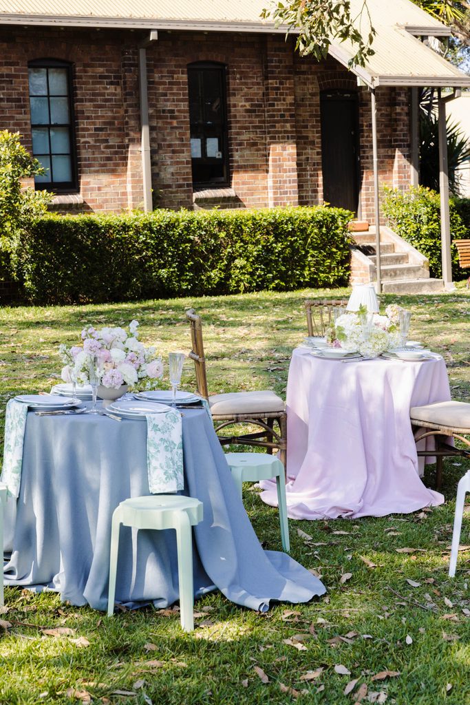 Emma Blomfield sits at one of two small, circular outdoor dining tables set with floral centerpieces and blue and pink Emma Blomfield rug collection linens on a lawn in front of a brick building.