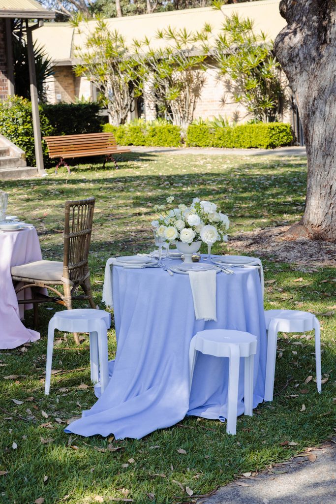 A round table with a blue tablecloth, white floral centerpiece, and four white stools set on an Emma Blomfield rug collection piece atop a lawn.