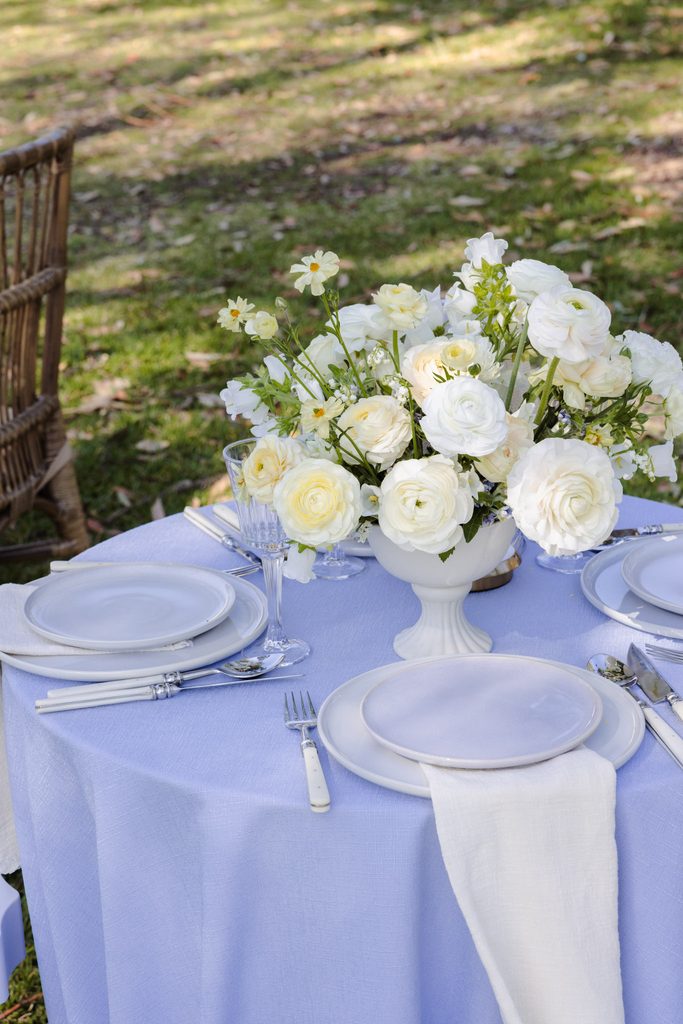 A round table covered by an Emma Blomfield rug collection piece and a light blue tablecloth, set for dining with a white ranunculus centerpiece.