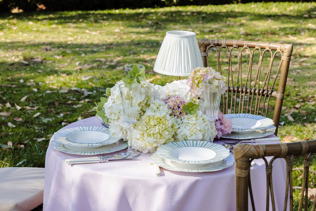 A round table set for two with plates, glasses, and a hydrangea centerpiece, placed on an Emma Blomfield rug on the lawn.
