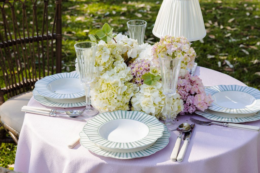 A round table set for a meal atop an Emma Blomfield rug features plates with green striped rims, crystal glassware, and a centerpiece of hydrangeas.