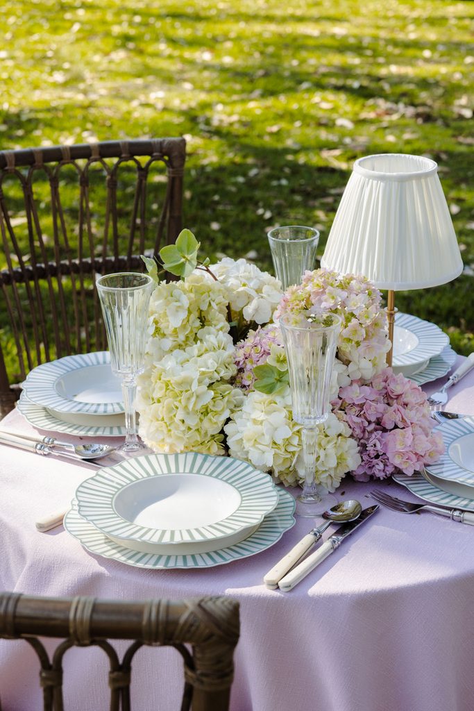 A round table set for an outdoor meal atop an Emma Blomfield rug, featuring place settings, floral centerpieces, and a small table lamp.