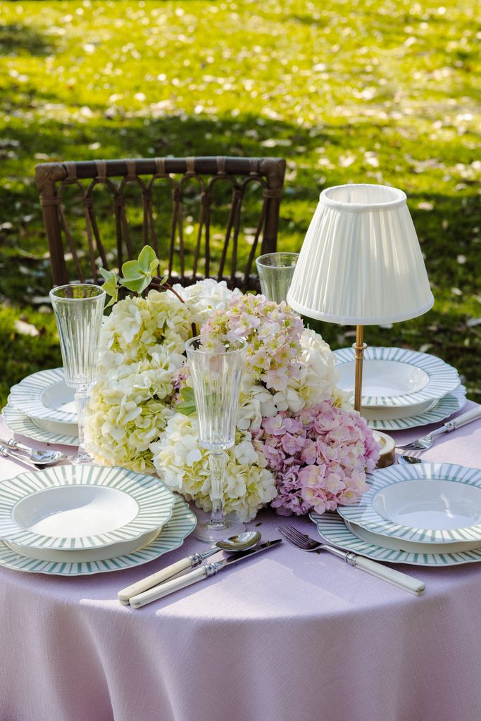 A round garden table set for two atop an Emma Blomfield rug features plates, cutlery, champagne flutes, a small lampshade, and a large hydrangea centerpiece.