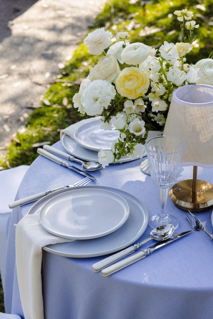 A table setting on an Emma Blomfield rug collection tablecloth features stacked plates, cutlery, a small lamp, and a floral centerpiece of white and yellow blooms.