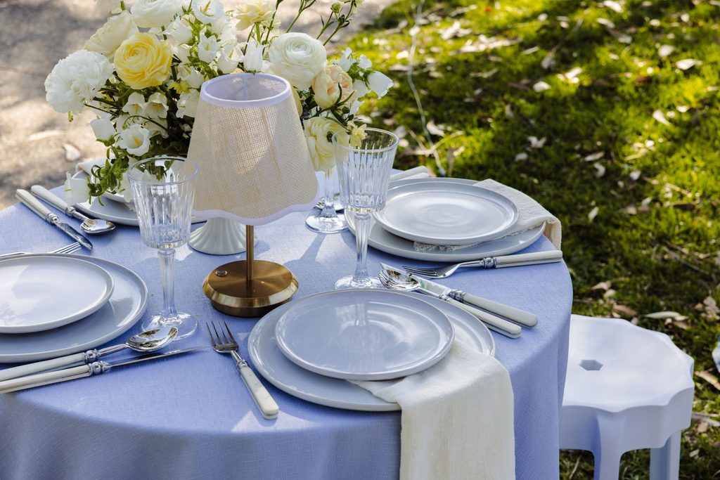 A round table set for dining atop an Emma Blomfield rug features a blue tablecloth, plates, silverware, glassware, a small lamp, and a floral centerpiece.