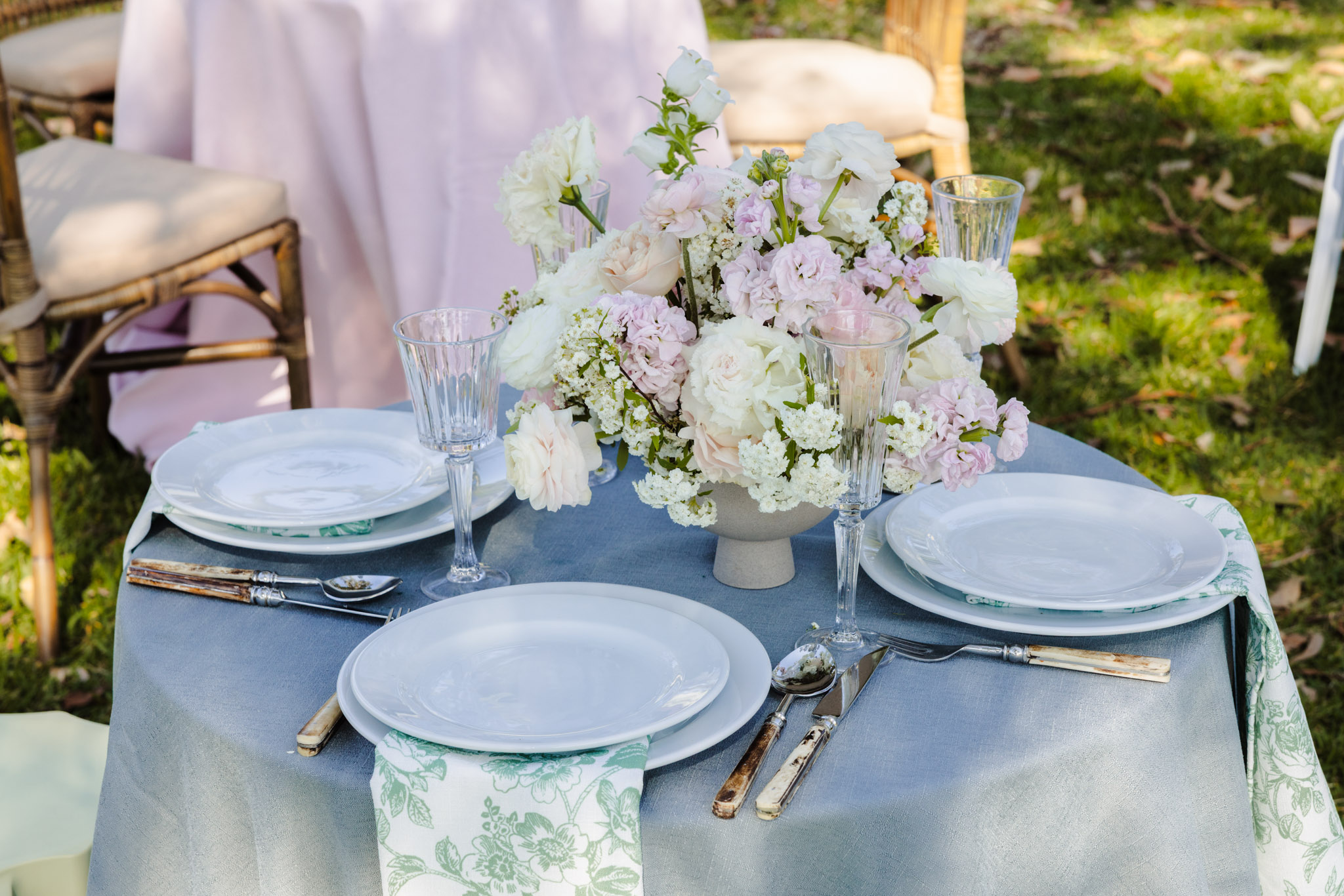 A round outdoor table set for three with white plates, glassware, and a floral centerpiece, situated atop an Emma Blomfield rug.