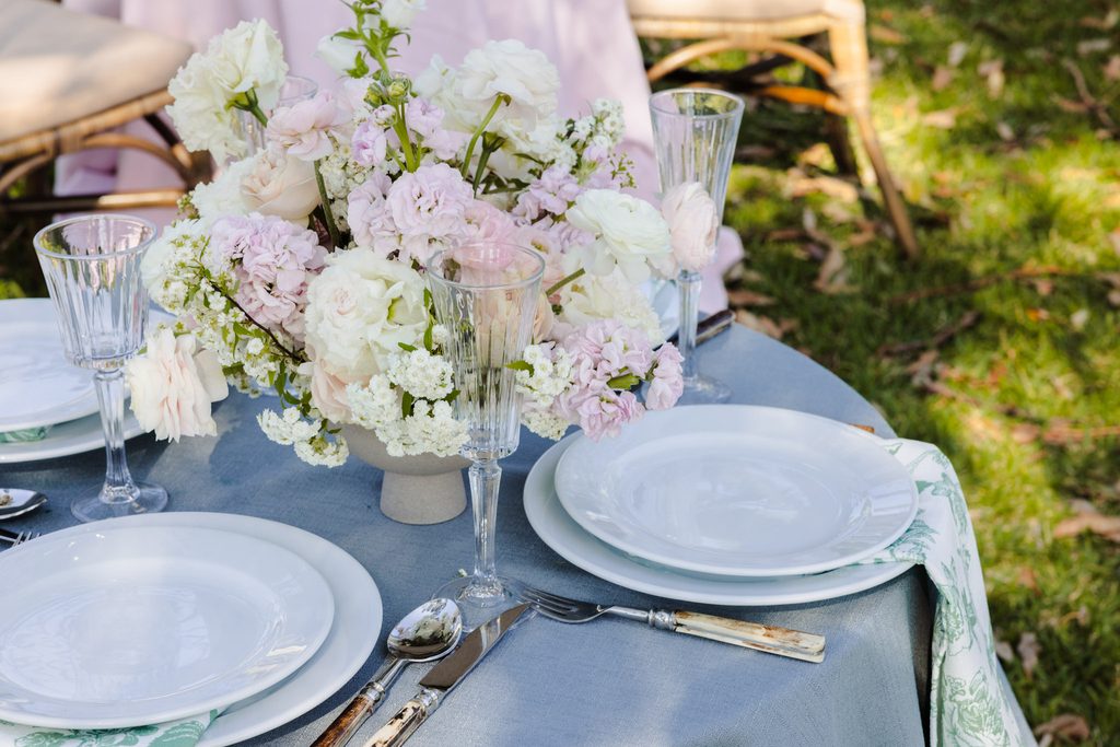 A round table set for an outdoor meal atop an Emma Blomfield rug, featuring white plates, glassware, and a central floral arrangement of white and pink flowers.