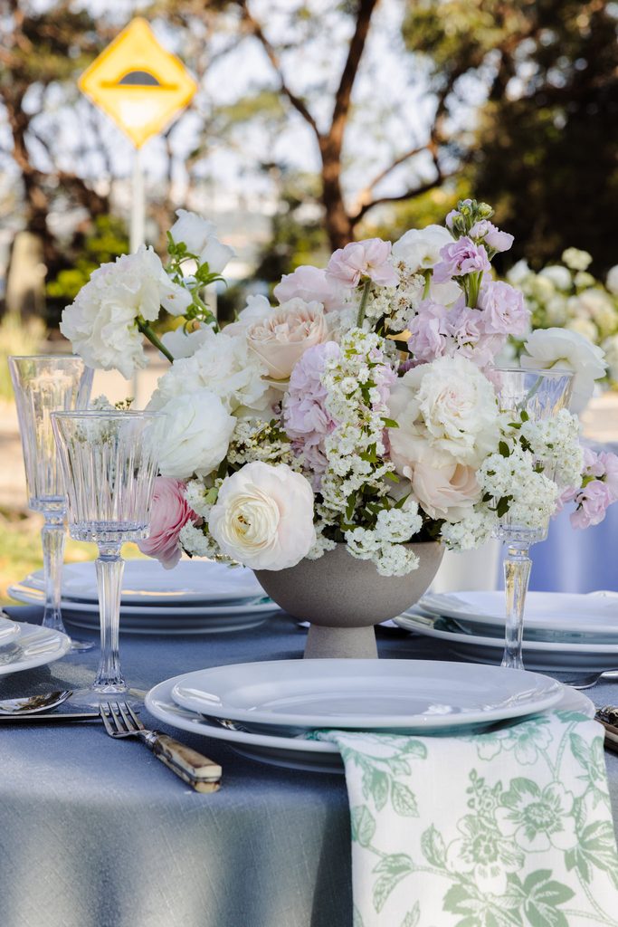 A large floral centerpiece of white and pink flowers in a grey bowl sits on an Emma Blomfield rug table setting, flanked by crystal glassware and white plates on a grey tablecloth.