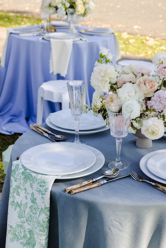 Round dining tables with floral centerpieces, white plates, glassware, and patterned napkins sit atop an Emma Blomfield rug at an outdoor event.