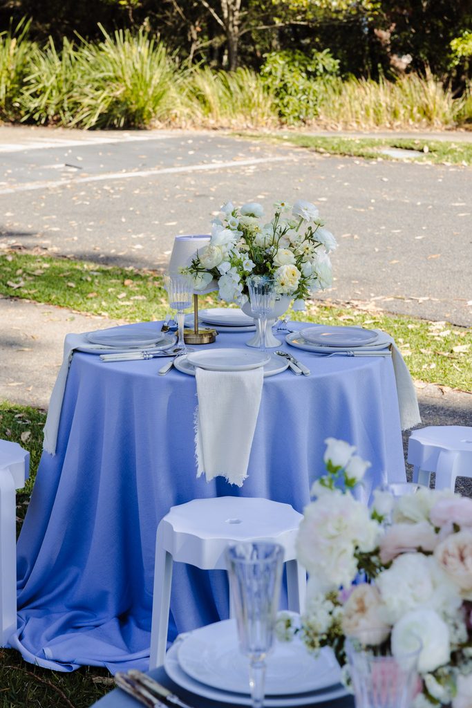 A round table covered in a periwinkle cloth sits atop an Emma Blomfield rug, set with plates, glassware, and a white floral centerpiece.
