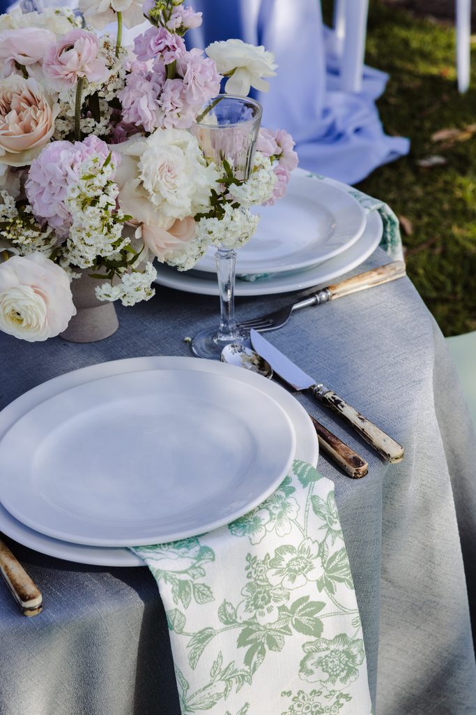 A place setting with white plates, a green floral napkin, vintage-style cutlery, and a floral centerpiece on an Emma Blomfield rug collection grey tablecloth.