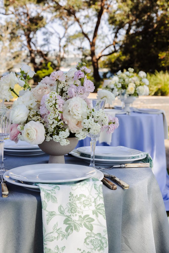 A floral centerpiece in a stone bowl sits on a gray Emma Blomfield rug table set with white plates and green patterned napkins.