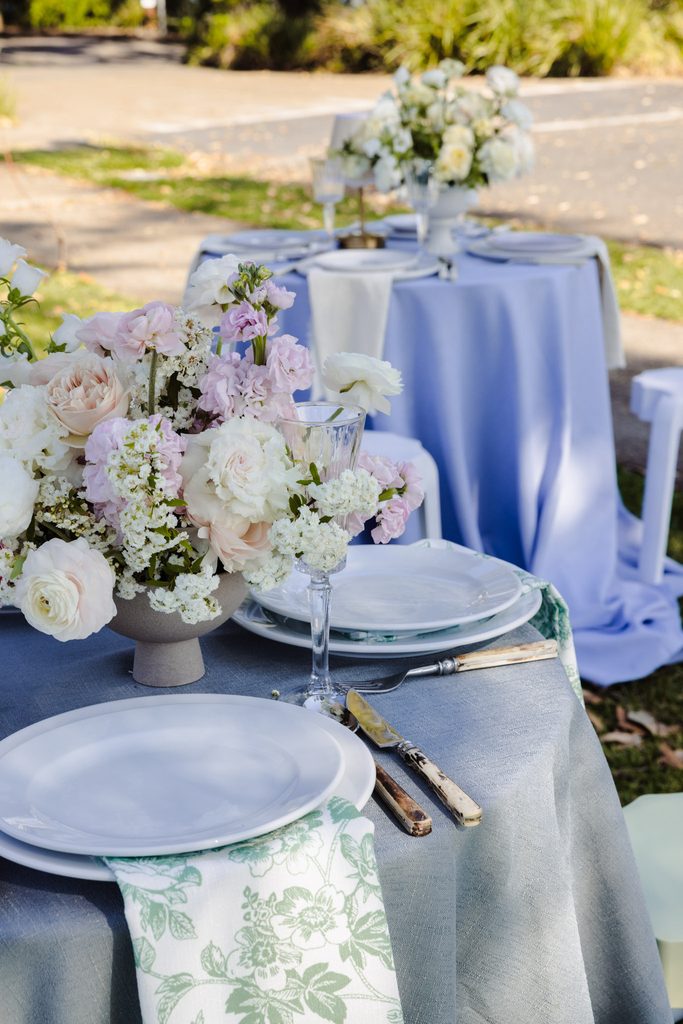Round tables set for an outdoor event atop an Emma Blomfield rug feature floral centerpieces, plates, and patterned napkins.
