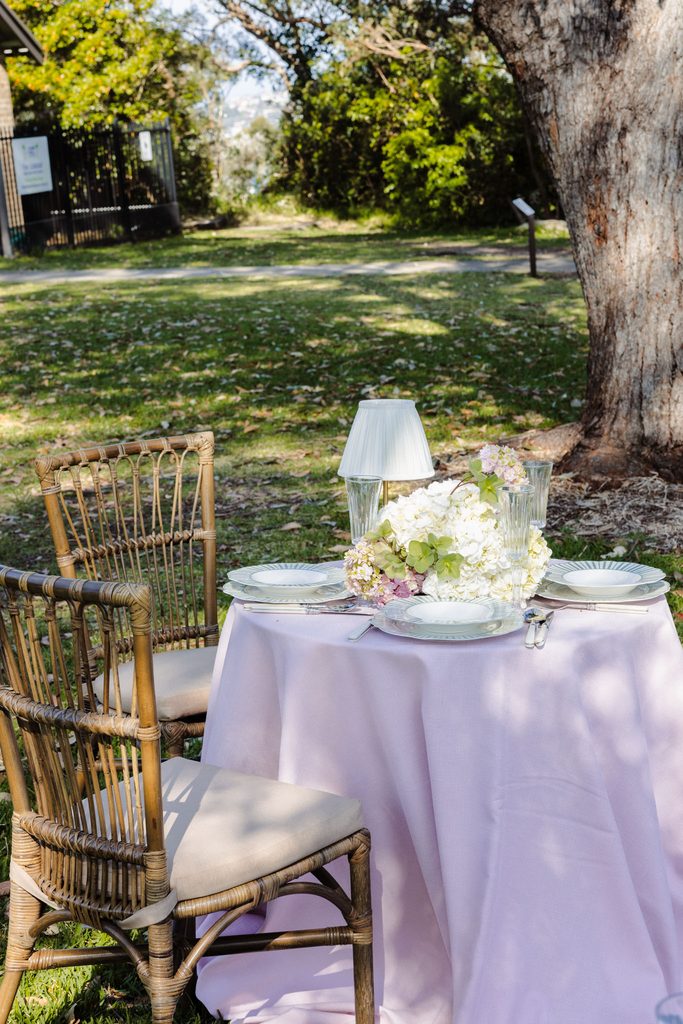 A table set for two with a pale pink tablecloth, floral centerpiece, and wicker chairs atop an Emma Blomfield rug on the lawn.