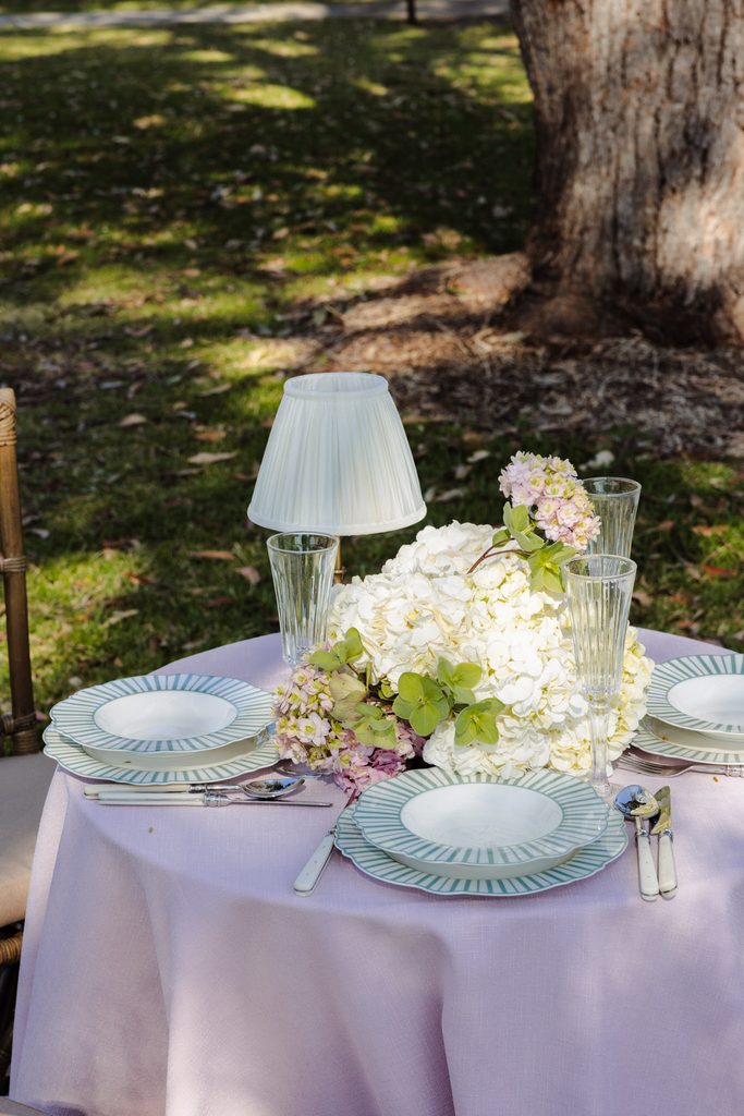 A round table with a pink tablecloth atop an Emma Blomfield rug is set with striped plates, glassware, a small lamp, and a centerpiece of white and pink hydrangeas.