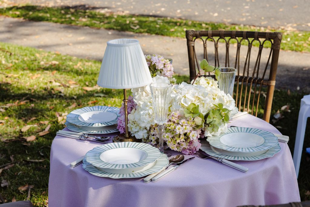 A small round table sits on an Emma Blomfield rug, set for two with pink linens, striped plates, glassware, a central flower arrangement, and a small table lamp.