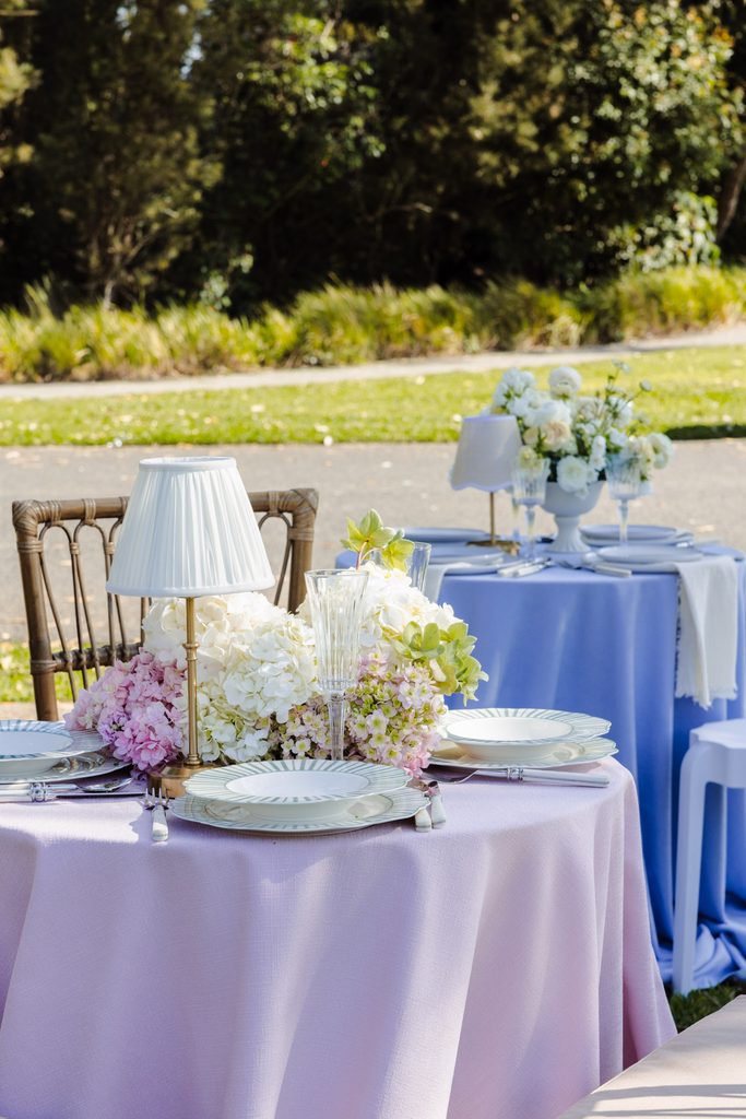 Emma Blomfield stands by round tables set for an outdoor event atop an Emma Blomfield rug, featuring floral centerpieces, small lamps, and place settings.