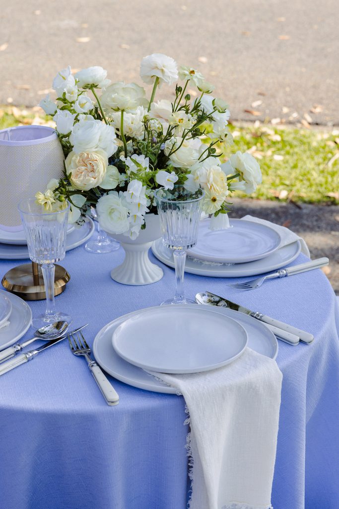 A round table with a blue tablecloth, white plates, silverware, crystal glasses, and a floral centerpiece of white flowers is set atop an Emma Blomfield rug.